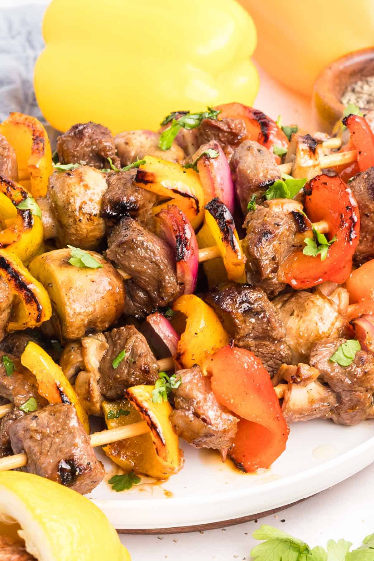 Chunks of sirloin are marinated then threaded onto skewers with onions, bell peppers, and mushrooms to make these beef kebabs. Side view of beef kebabs on a white plate