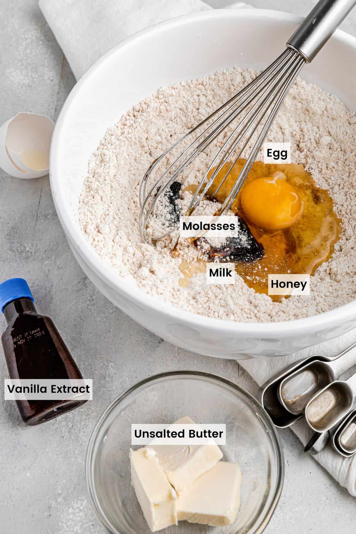 Eggs and other wet ingredients on top of the dry ingredients in a bowl with butter pieces and a bottle of vanilla next to it with text labels.
