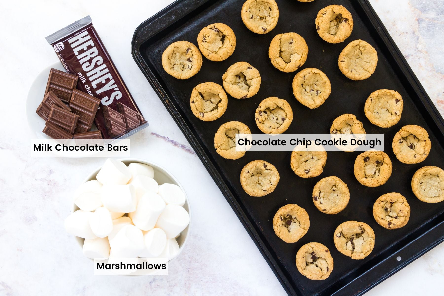 S'Mores Cookie Cups combine the warm, chocolate-studded charm of the classic chocolate chip cookie with a melty, sticky campfire classic. A pan of fresh-baked chocolate chip cookies beside a bowl full of chocolate squares and a second bowl of marshmallows with text labels.