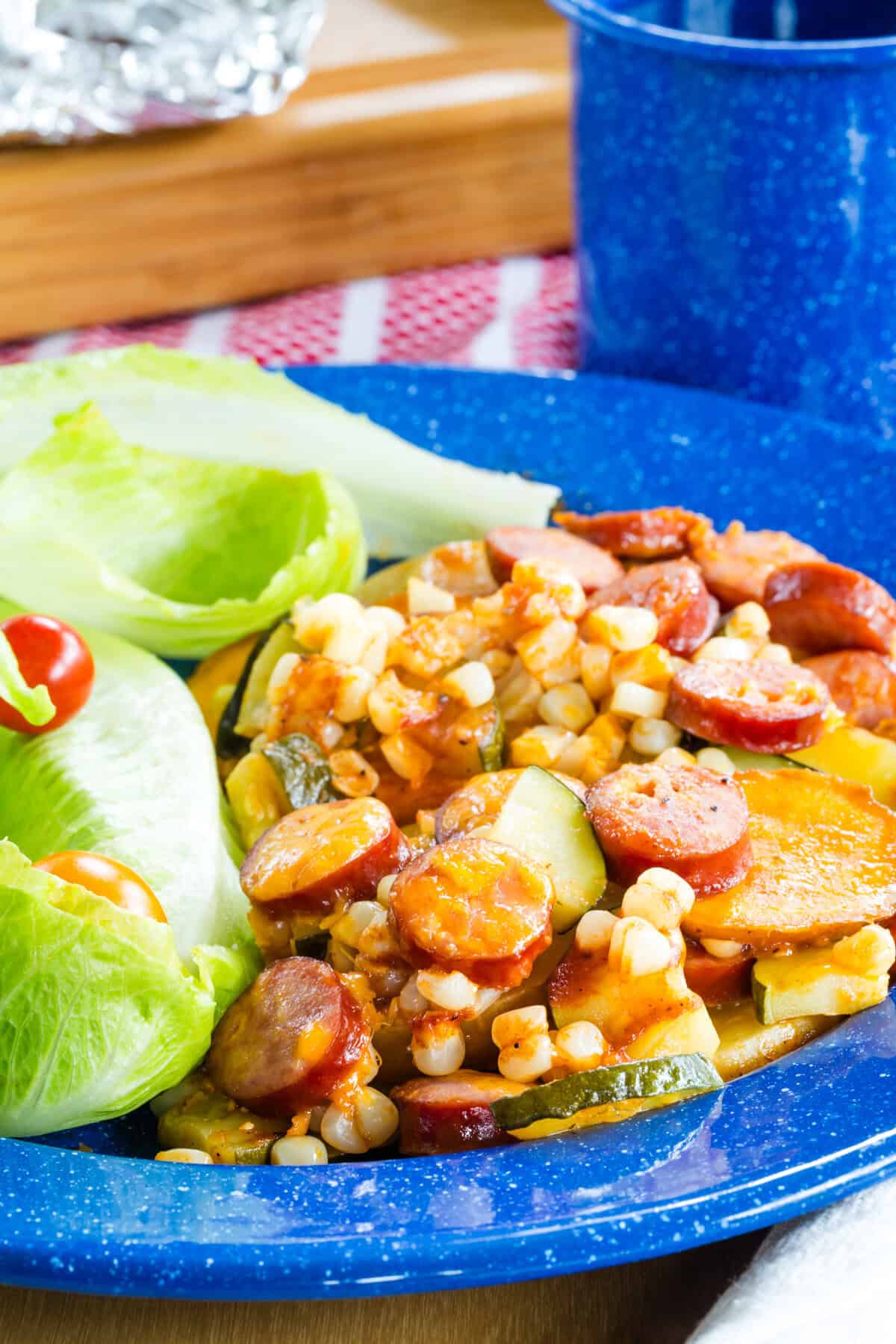 Sausage, corn, potatoes, and zucchini coated in barbecue sauce and melted cheese on a plate with lettuce.