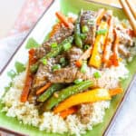 Beef stir fry with carrots, peppers, and green beans over cauliflower rive on a green rectangular plate.