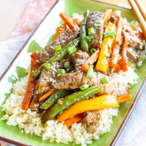 Beef stir fry with carrots, peppers, and green beans over cauliflower rive on a green rectangular plate.