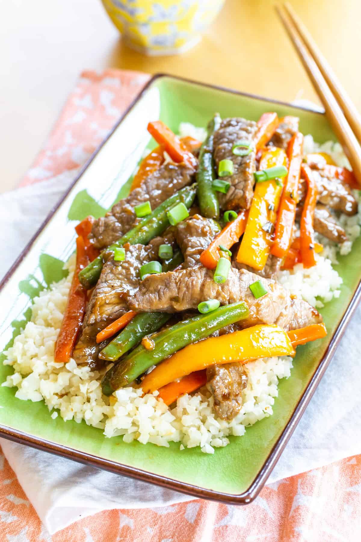Beef stir fry with carrots, peppers, and green beans over cauliflower rive on a green rectangular plate.