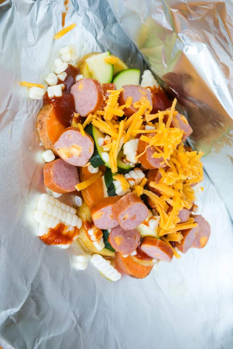 Uncooked corn, zucchini, and potatoes with sausage slices and shredded cheese on a piece of aluminum foil.