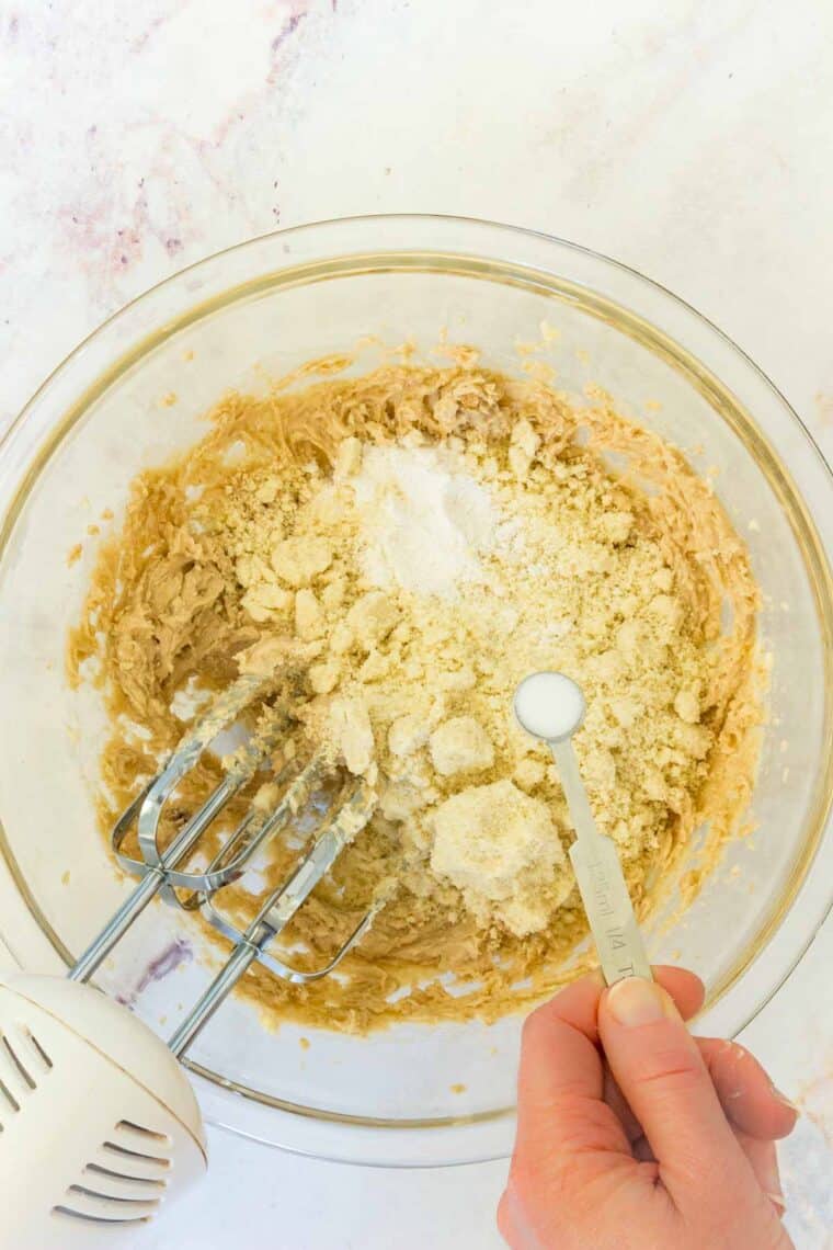 An easy recipe for edible chocolate chip cookie dough that's gluten-free and 100% safe to eat by the spoonful! The perfect sugary treat. Salt being sprinkled on top of a bowl with creamed butter and sugar, flour, and almond flour in it.