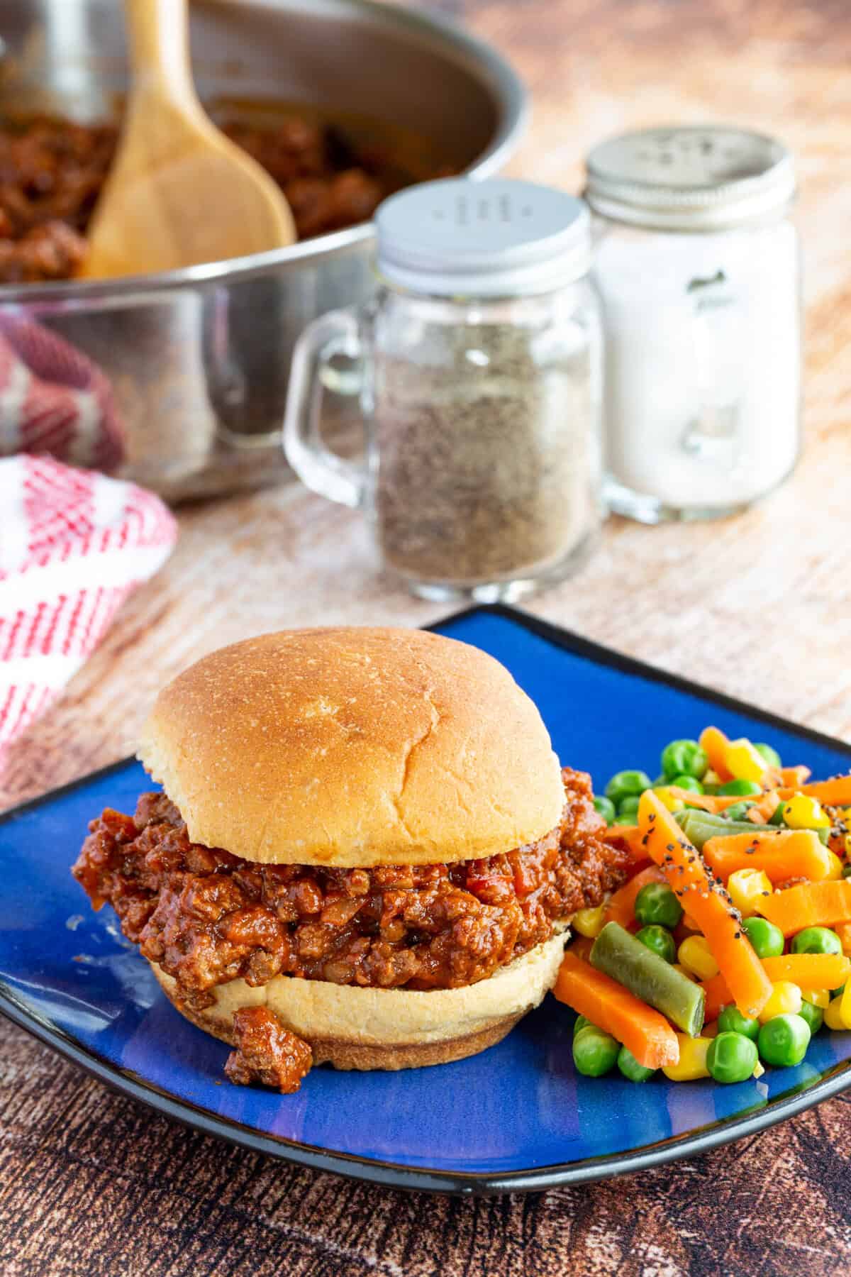 Sloppy joes on a bun with mixed veggies on a blue plate.