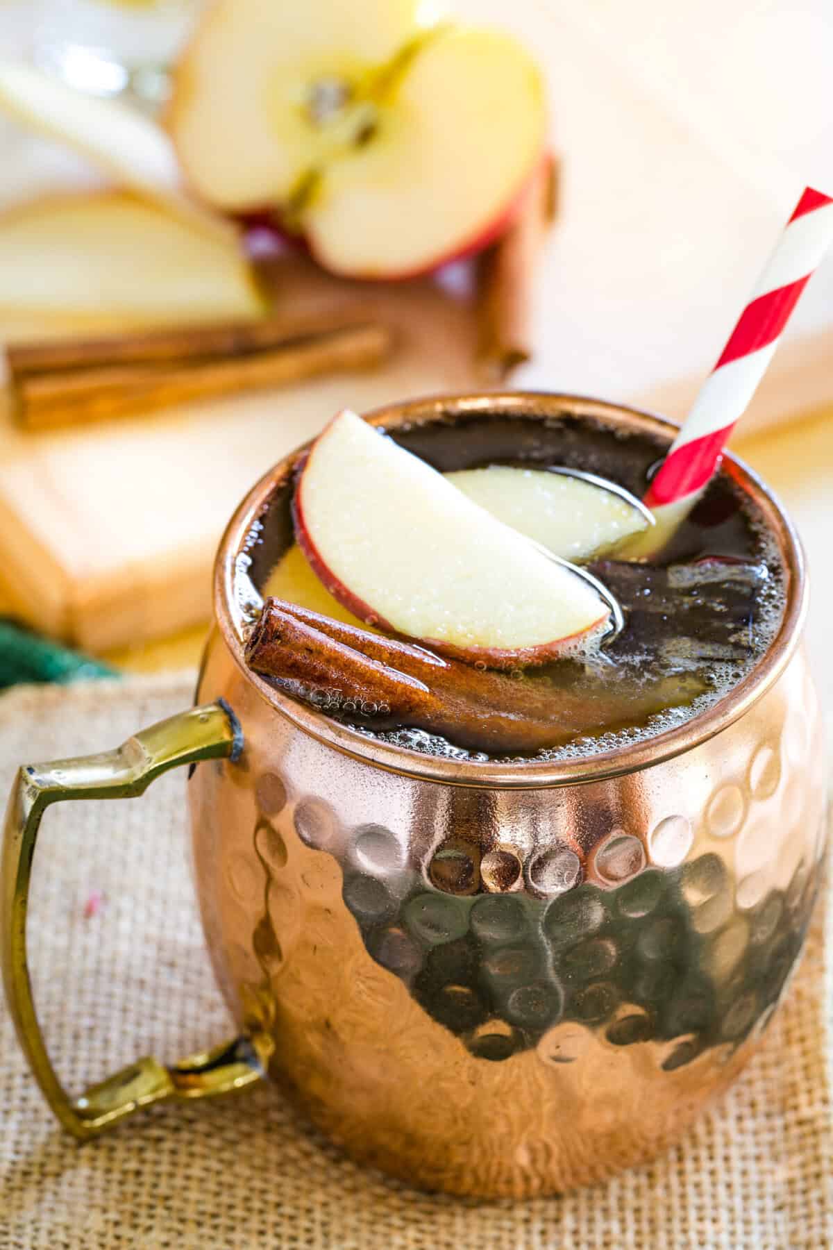A cocktail garnished with apple slices and a cinnamon stick in a copper mug.