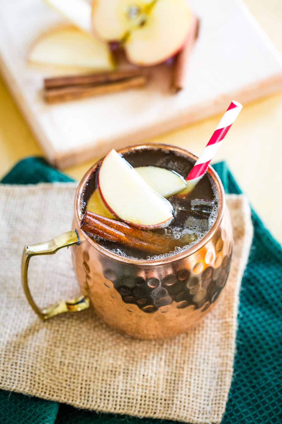 An apple cider cocktail in a copper mug next to a cutting board with aple slices.