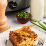 A slice of crockpot breakfast casserole on a plate with a fork in front of the crockpot