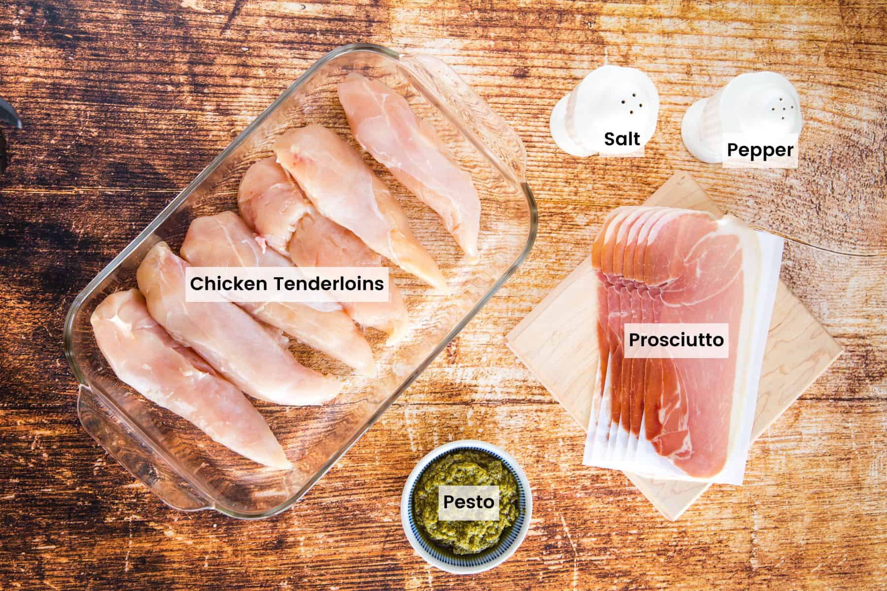 Chicken tenderloins, prosciutto, a small dish of pesto, and salt and pepper shakers on a wooden tabletop.