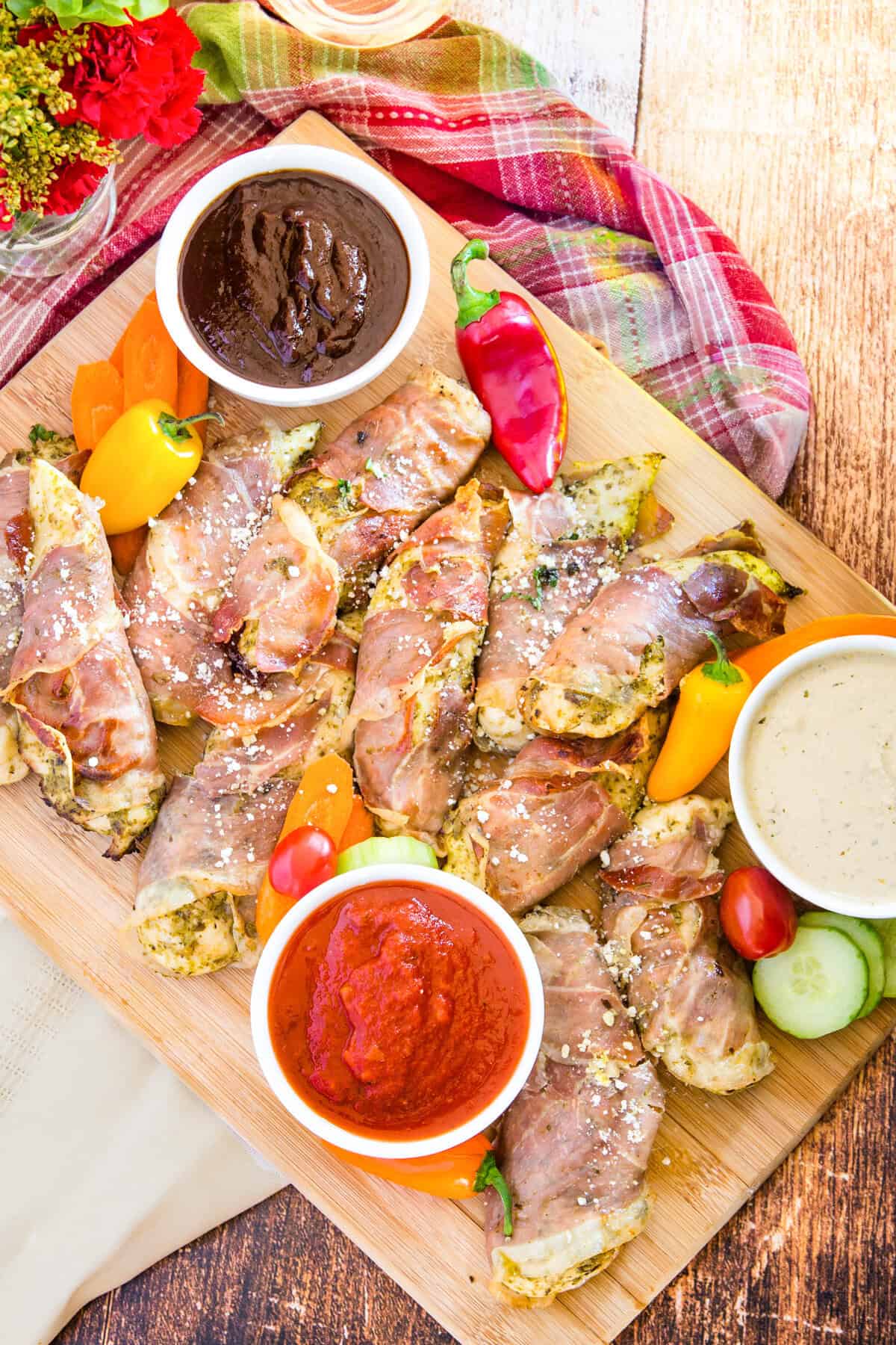 Looking down on a wooden board with prosciutto-wrapped chicken and vegetables on it with bowls of dips and sauces.
