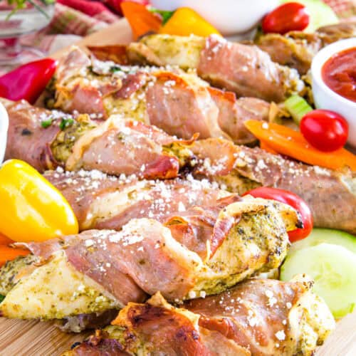 Prosciutto chicken tenders piled on a cutting board with vegetables.