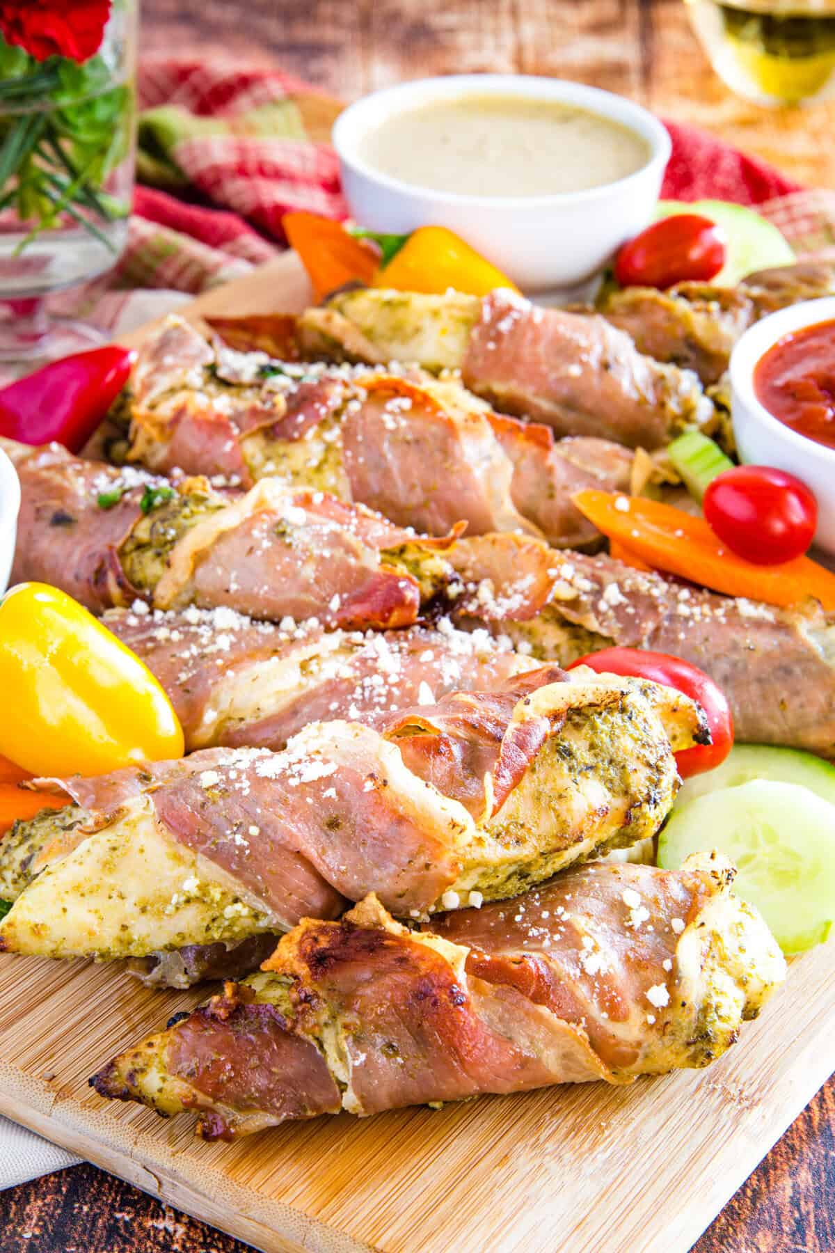 Prosciutto chicken tenders piled on a cutting board with vegetables.