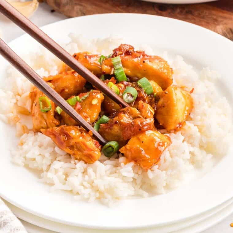Chopsticks picking up a piece of Sweet and Spicy Chicken from on top of a bed of rice.